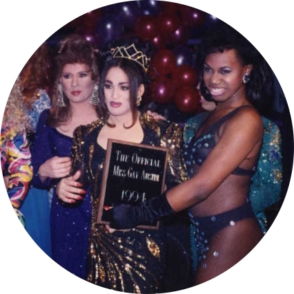 A group of drag performers in elaborate clothing surround the winner of the Miss Gay Texas America pageant, who is being handed a plaque.