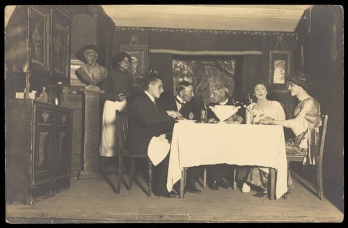 Download the full-sized image of Belgian prisoners of war, some in drag, sit at a table within a very detailed stage set at Friedberg camp, Germany. Photographic postcard, 191-.