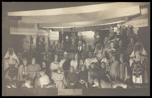 Download the full-sized image of Amateur actors, some in drag, pose on a crowded stage. Photographic postcard, 191-.