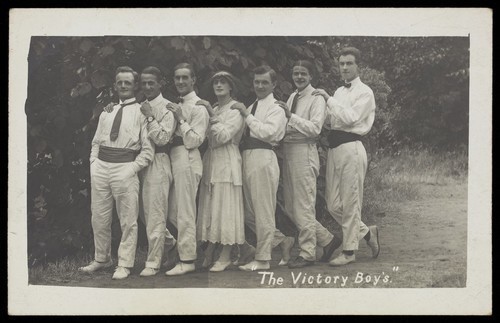 Download the full-sized image of Entertainers, one in drag, pose in a line in front of foliage. Photographic postcard, 191-.